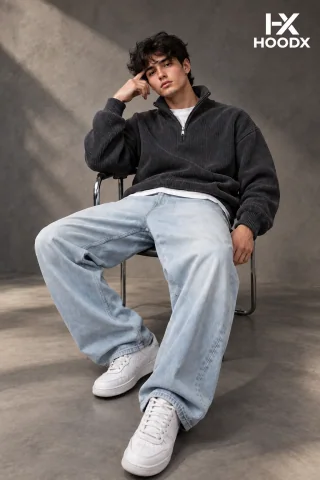 Seated model wearing a black HoodX zip top, relaxed grey trousers, and white designer trainers.