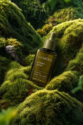 Lumera Vitamin C serum glass dropper bottle resting on natural moss-covered rocks in soft sunlight.